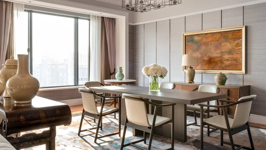 A sophisticated dining room with a rectangular wooden table, cream-coloured chairs, decorative vases and large window. A framed painting and lamp adorn the sideboard.