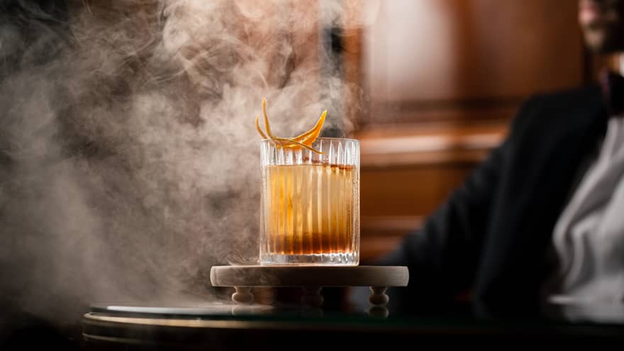Smoke billows in the air as a glass dome is being lifted off of a whiskey-based cocktail in a rocks glass garnished with orange peel