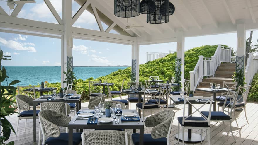 Covered restaurant patio with vasious square tables each set with four blue-and-white chairs with a view of the ocean in the background