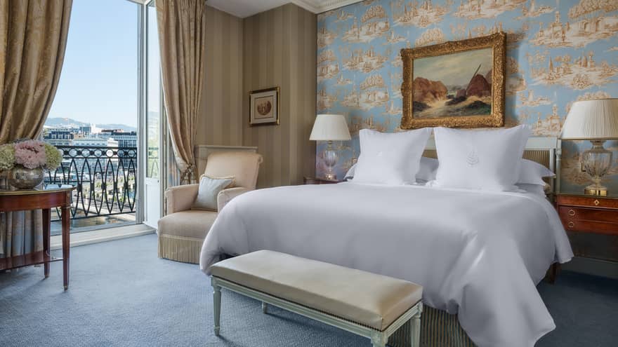 Luxury hotel room with a king bed, nightstands with lamps, bench, patterned wallpaper, armchair, framed artwork and a balcony with city view
