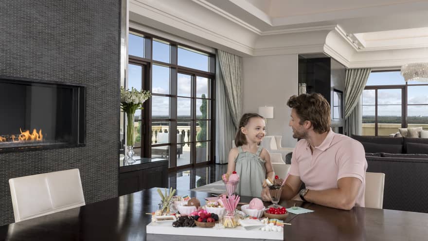 Man and young girl sit at dining table in hotel suite, next to fireplace