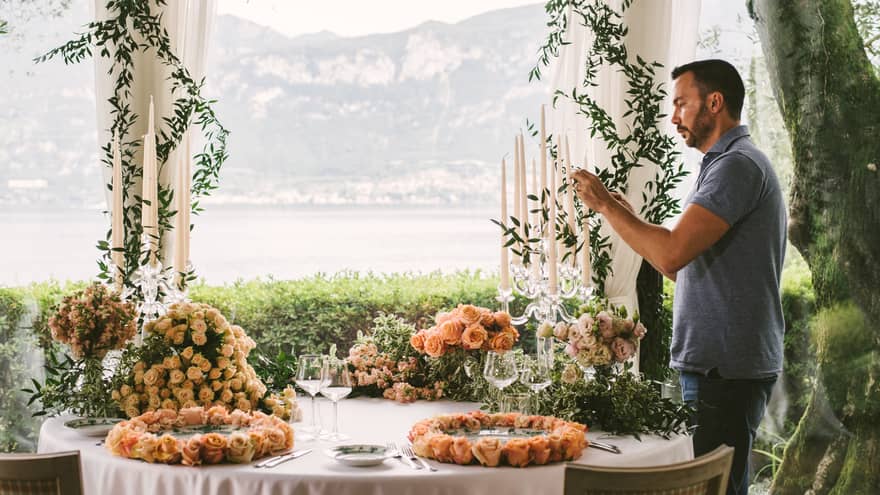 Man arranges tall candles in candelabra at table with pink rose floral arrangements, water views