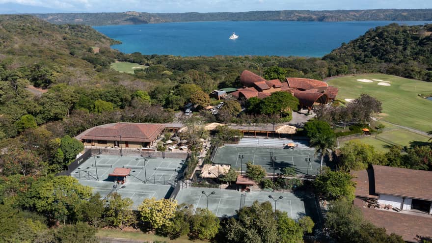 Aerial view of resort tennis centre, surrounded by trees with the ocean in the near distance