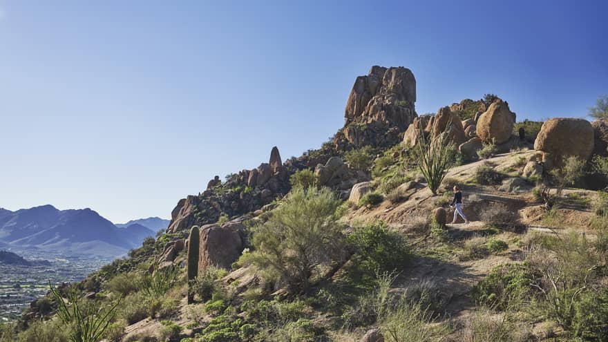 Woman walks along trail on rocky desert mountain under blue sky
