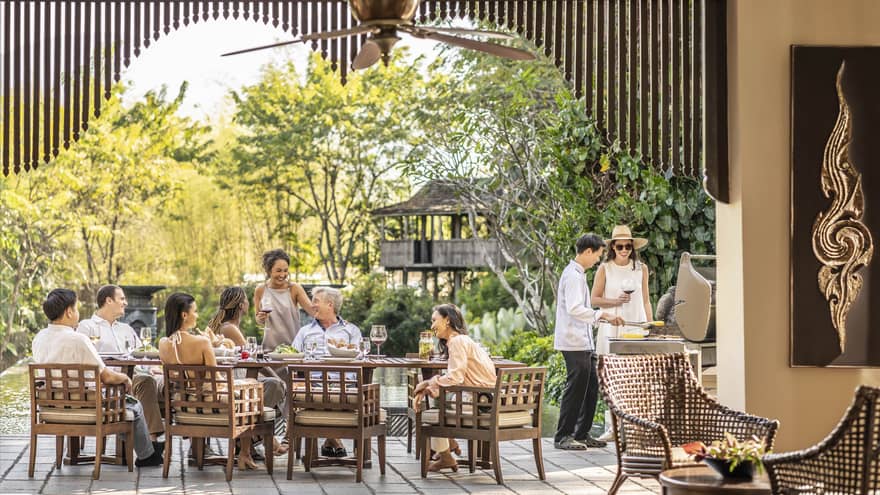 A long brown table with a family sat at it, in front of large trees.