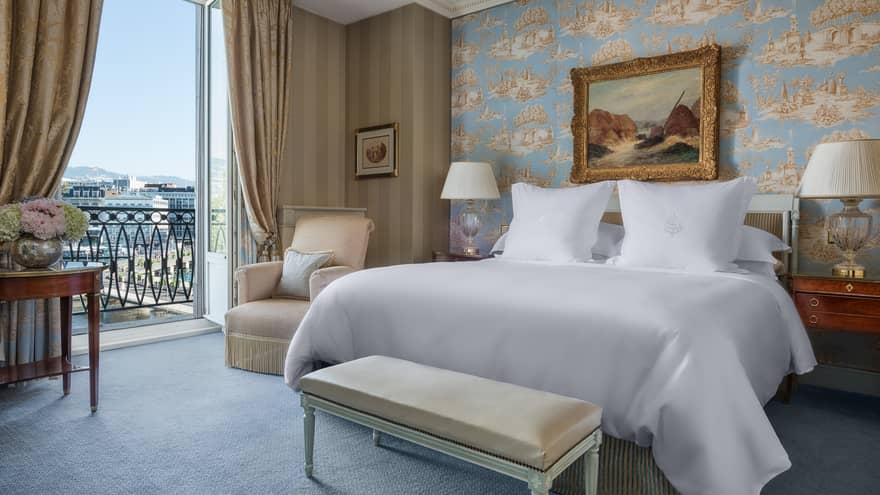 Luxury hotel room with a king bed, nightstands with lamps, bench, patterned wallpaper, armchair, framed artwork and a balcony with city view