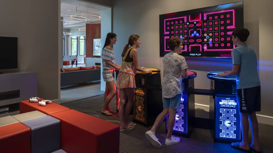Four smiling teenagers stand at multi-coloured video-game pedestals facing a large TV screen in a dimly-lit games room.