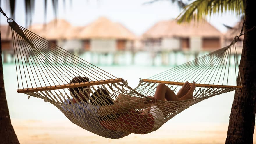 Couple lies in rope hammock between two palm trees, with beach, ocean and overwater bungalows in background