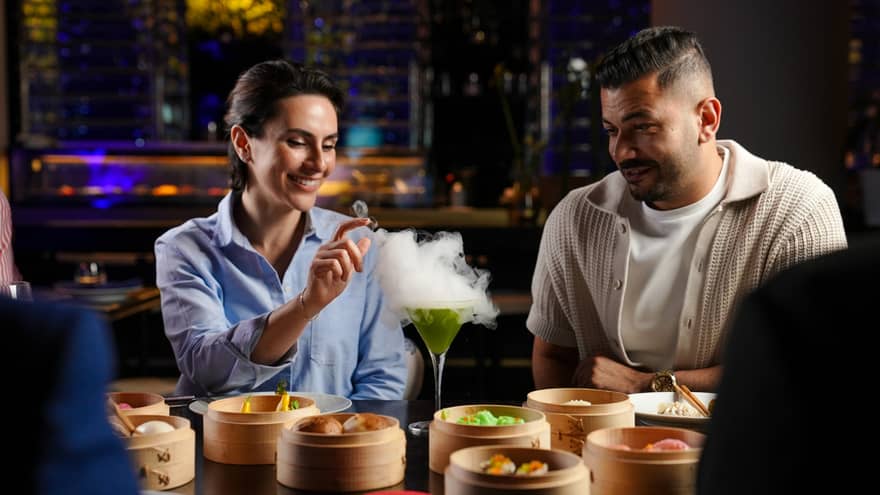 Two people sit at a table filled with baskets of dim sum and a green cocktail with a plume of smoke floating up from the cocktail