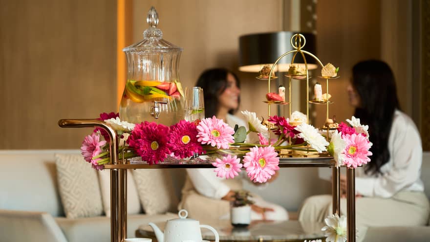 Bar cart decorated with pink Gerber daises carries a gold tray of pastries, a white tea pot and two mugs, and a large glass carafe of fruit-infused water