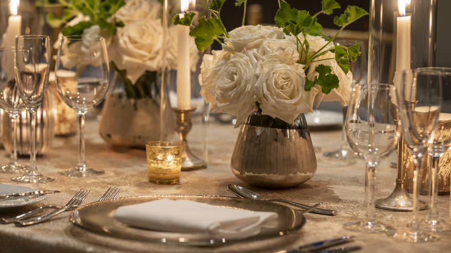 A dining table with white flower center pieces, silver plates, and wine glasses.