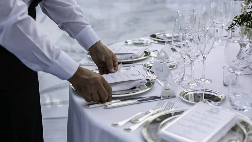 A server in a white shirt sets a formal dining table, placing a menu on a silver plate. The table is elegantly arranged with glassware, silverware and name cards.