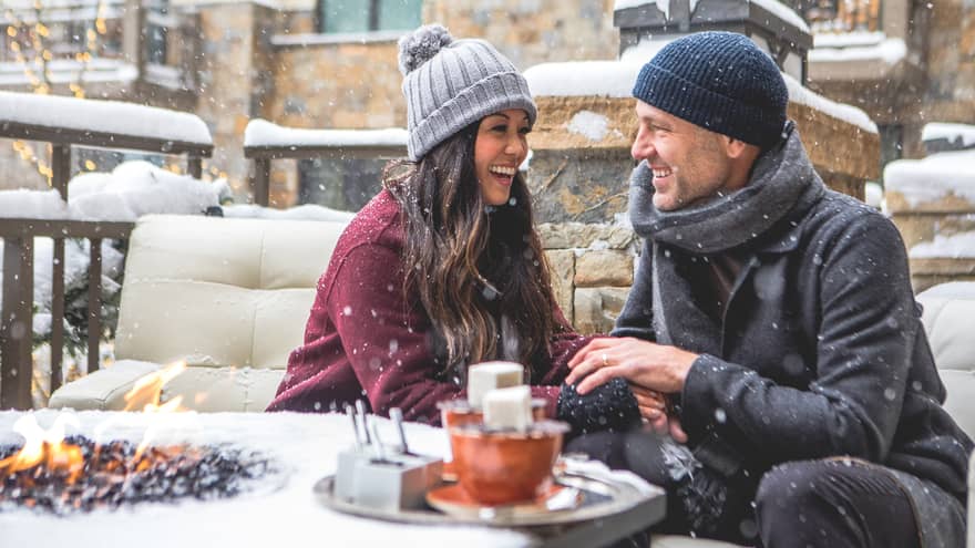 Smiling couple sitting by a firepit in a snowy outdoor setting, bundled in winter coats and hats. Snowflakes fall around them as they enjoy a cosy moment. 