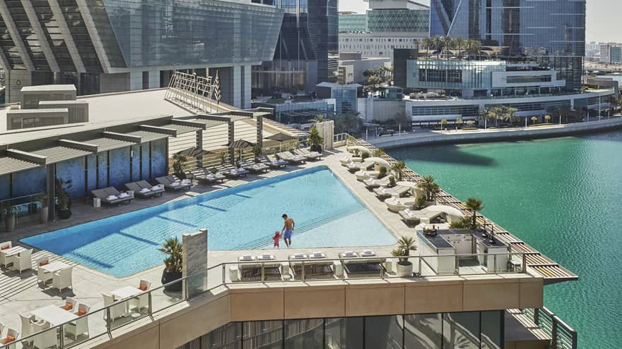A wide shot of a man and child walking around a triangular pool overlooking the water and Abu Dhabi skyscrapers in the background. 