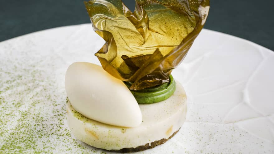 Structural cream-colored dessert with a dollop of green and a glass-like sheet of gold