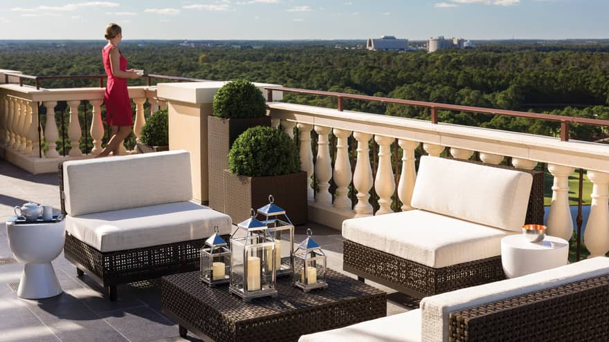 Woman stands at edge of sunny rooftop balcony near plush white patio furniture
