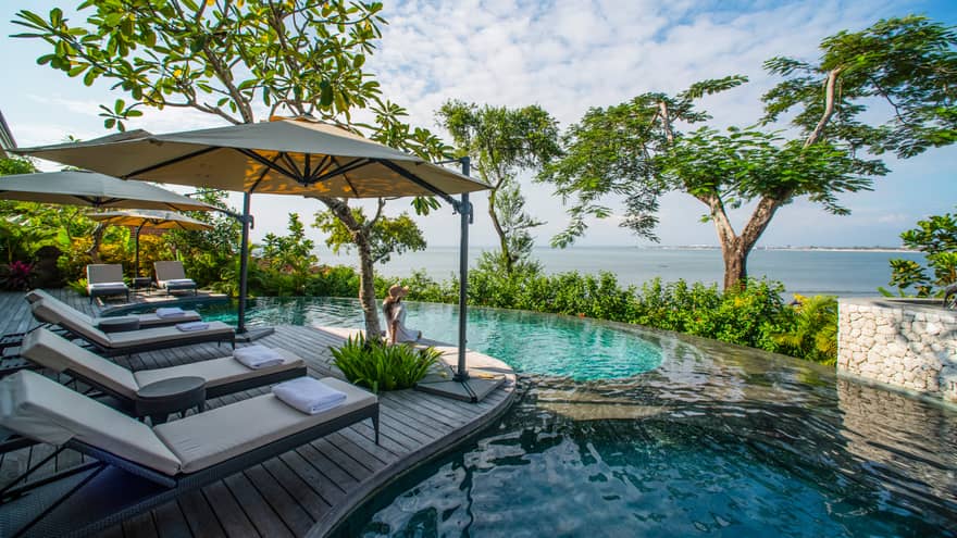 Lounge chairs with umbrella on wooden deck overlooking infinity pool, trees and ocean