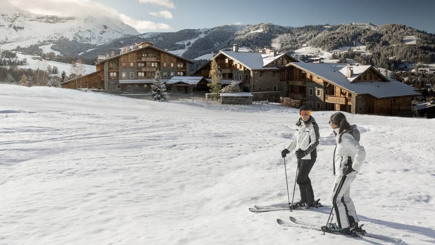 Man, woman on skis at top of snowy hill, Four Seasons Hotel Megeve in background