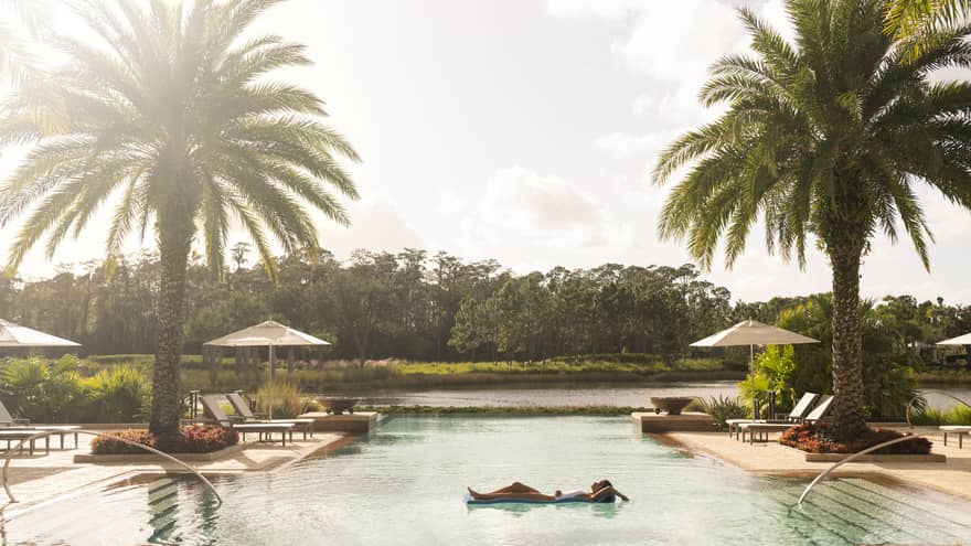  A serene pool scene at Four Seasons Resort Orlando, with a person floating on a pool float, palm trees, sun umbrellas, and a lush backdrop.