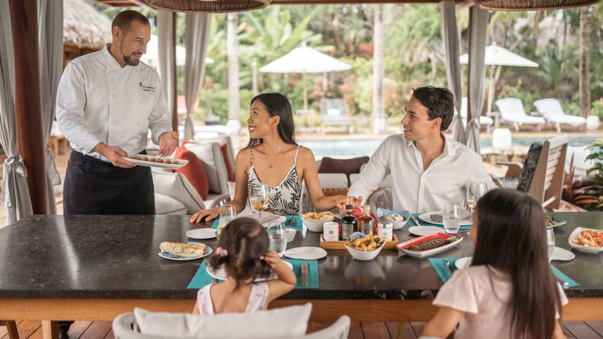 Chef serving a family seated at an outdoor dining table, with various dishes. The setting includes tropical décor, a poolside view and relaxed ambiance. 