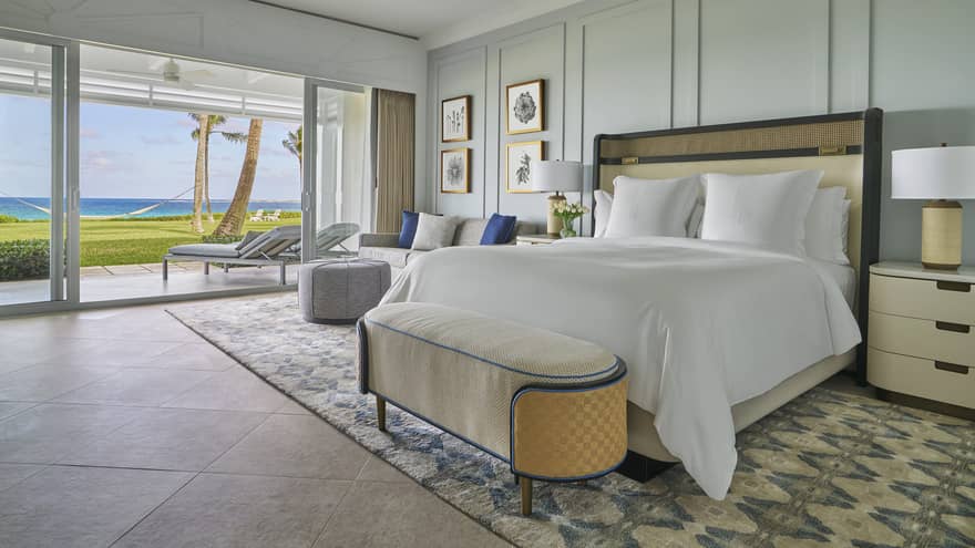 Luxury oceanfront bedroom with a large bed, cozy seating and sliding glass doors opening to a private patio with lounge chairs 