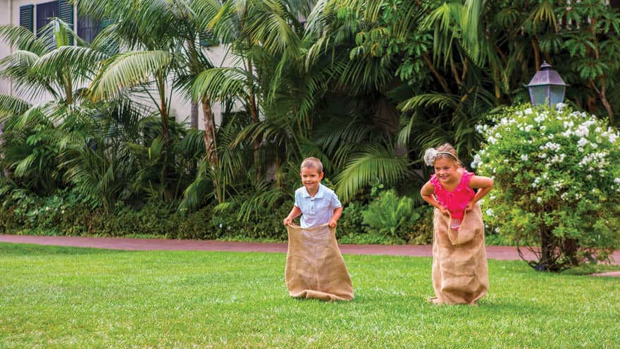 Two smiling children race across lawn in burlap sacks