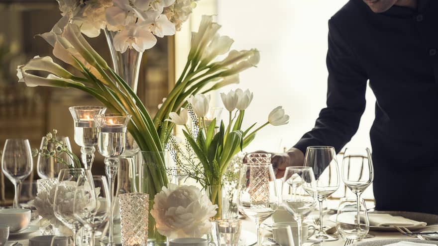 Hotel staff sets elegant banquet dining table with crystal glasses, vases with white flowers