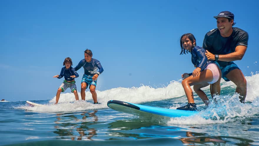 A pair of smiling adults and children standing on two surfboards, wearing swimsuits and rash guards, riding a small wave.