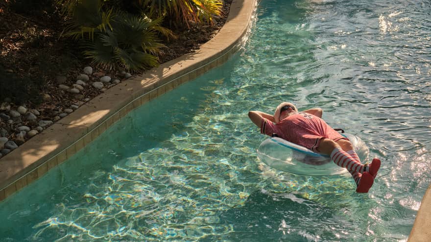 Santa wears his red hat, red short-sleeve onsie and red-and-white striped socks while floating on a clear inner tube on a lazy river