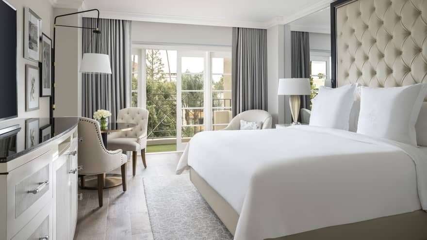 A hotel room with a large bed with white sheets, black counter on white dresser, three chairs, and a large window.