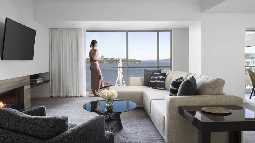 Woman stands at sunny window near L-shaped sofa in Deluxe Elliot Bay Suite 