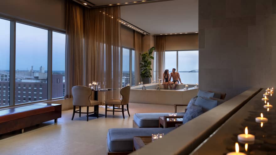 A couple enjoying a candlelit hot tub in a luxury hotel spa, with elegant seating, soft lighting and large windows offering a view of a distant bridge
