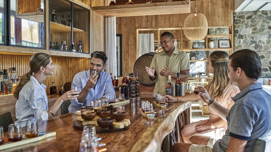 Four people sit around a live-edge wooden counter filled with selections of rum and chocolates as a rum master explains the flavors
