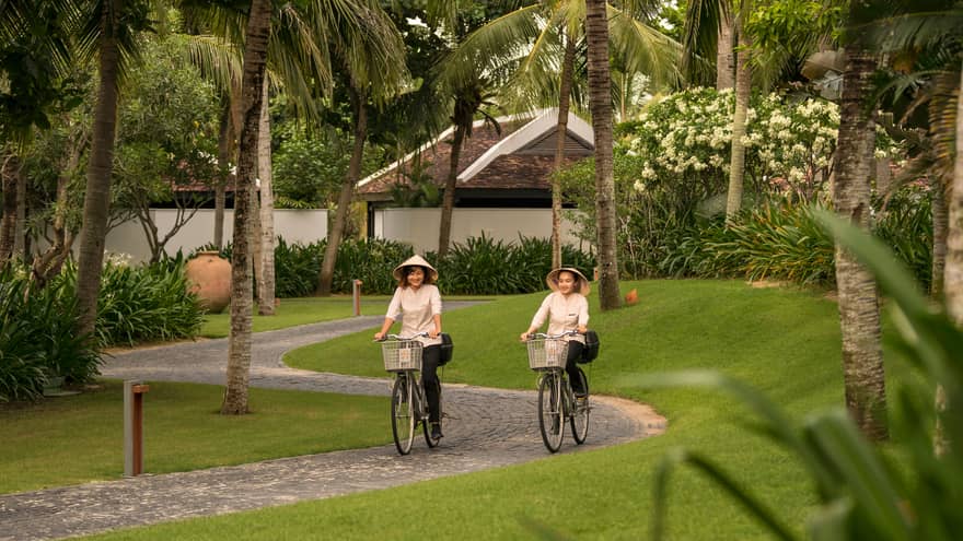 Two women wearing sun hats ride bicycles down winding brick path on resort 
