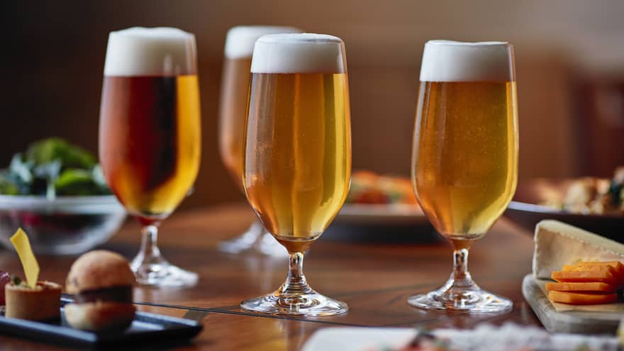 Four tall beer glasses filled with beer on a table with cheese, crackers and appetizers