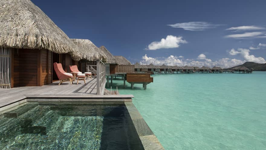 Overwater bungalows with thatched roofs, private pools and lounge chairs overlooking a clear turquoise lagoon under a bright blue sky.
