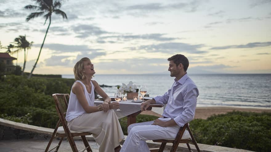 Two people enjoying wine outdoors at a beachfront table with ocean view at sunset