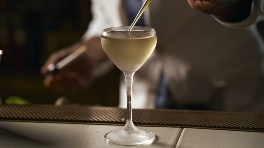 A hand uses a pipette to top up a gold-coloured martini in a stemmed glass with a rounded edge, in a darkly lit room.
