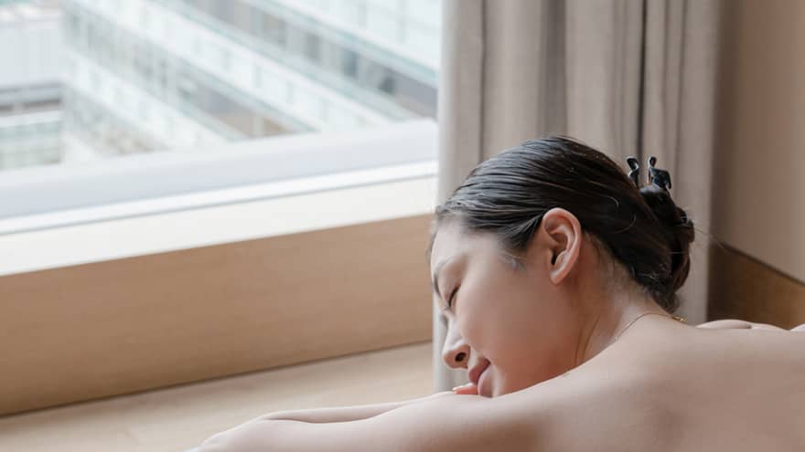 Person relaxing on massage table by a window with eyes closed, enjoying a peaceful moment