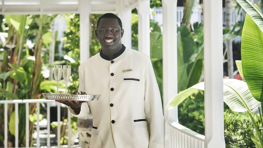 Man standing outdoors near plants and trees holding tray with two glasses of champagne.