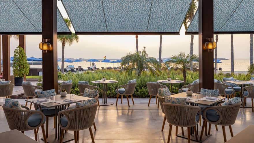 Covered restaurant terrace with grey and blue tables decor leads out to an open-air deck with greenery and the beach in the distance