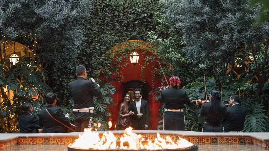 Two guests watch musicians play in a greenery-filled courtyard, with a fountain with a flame feature in the foreground.