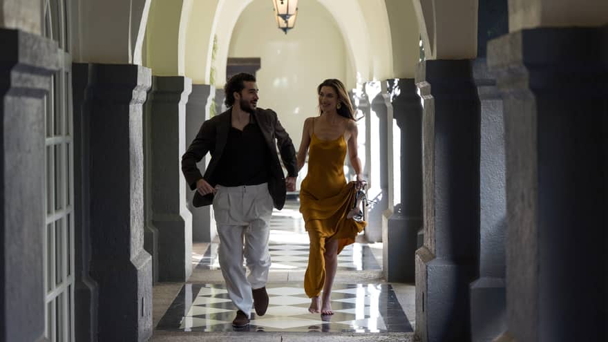 Two people walk down an arched hallway with a black-and-white tiled floor.