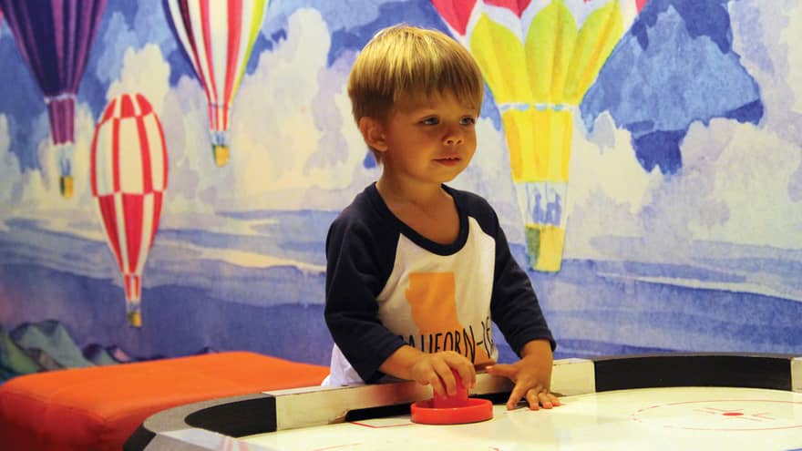 Young boy plays air hockey in Kids Activity Room in front of colourful mural of hot air balloons