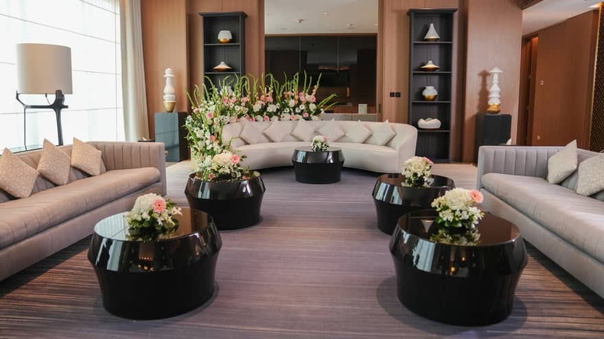 Hotel suite living room furnished with three light-grey curved sofas all facing each other and five round black coffee tables each topped with flowers