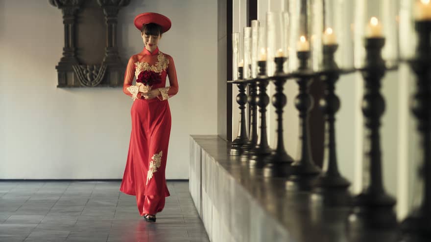 A person in a traditional red Vietnamese dress, carrying a bouquet, walking gracefully along a hallway lined with candles.