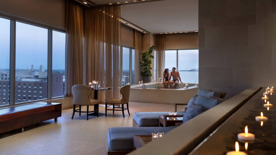 A couple enjoying a candlelit hot tub in a luxury hotel spa, with elegant seating, soft lighting and large windows offering a view of a distant bridge