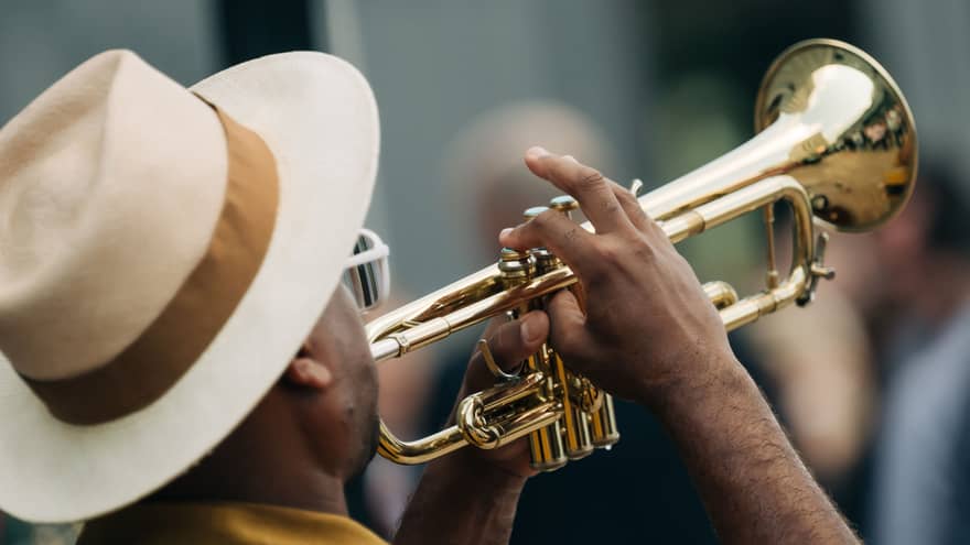 A man wearing a cream-and-taupe fedora with a brown ribbon plays a trumpet