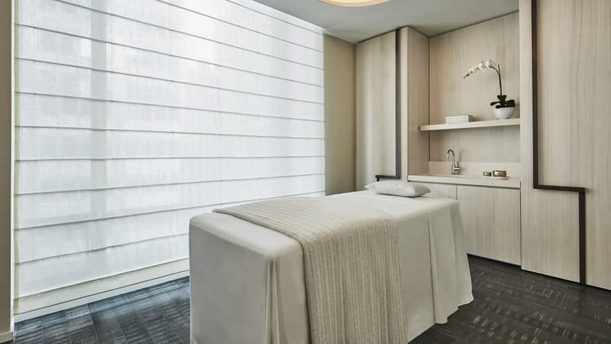 Five-star luxury hotel spa treatment room with a massage table, large sheer window blinds, soft lighting and a shelf with an orchid