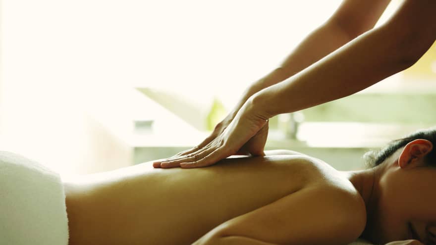 Masseuse reaches over, massages woman's bare back as she lies on spa table